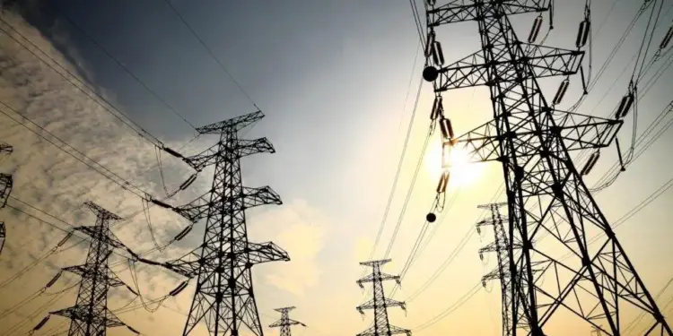 Torres metálicas de alta tensión con cables de transmisión contra un cielo nublado y luz solar.