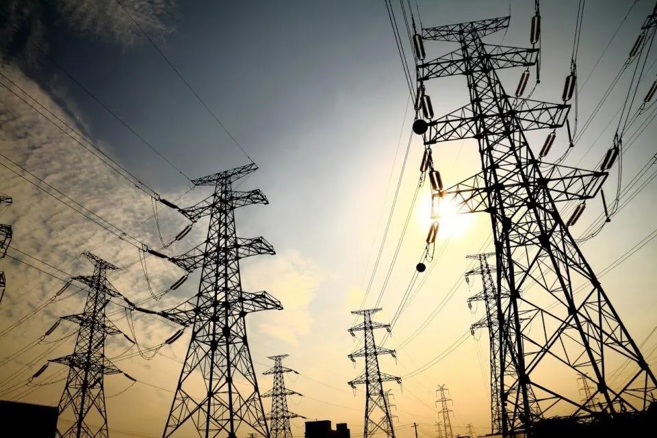 Torres metálicas de alta tensión con cables de transmisión contra un cielo nublado y luz solar.