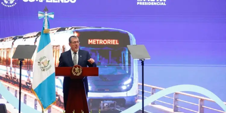 Hombre con traje hablando en un atril frente a una bandera y una pantalla con la imagen de un tren llamado Metroriel.