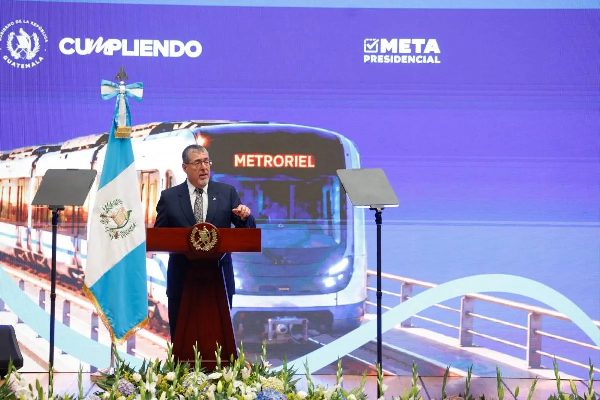 Hombre con traje hablando en un atril frente a una bandera y una pantalla con la imagen de un tren llamado Metroriel.