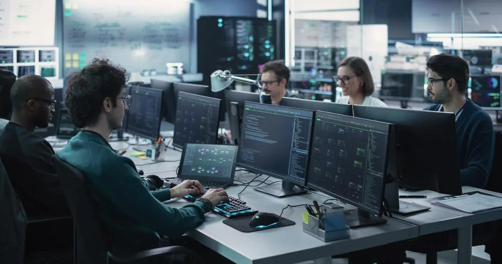 Grupo diverso de profesionales de TI trabajando en computadoras con múltiples monitores mostrando código y datos en una oficina moderna. Ambiente de desarrollo tecnológico y análisis.
