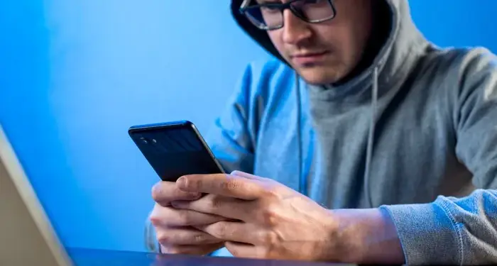 Hombre con sudadera y gafas usando un teléfono móvil junto a una laptop sobre fondo azul.