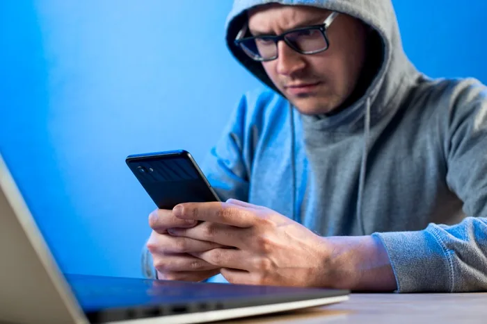 Hombre con sudadera y gafas usando un teléfono móvil junto a una laptop sobre fondo azul.