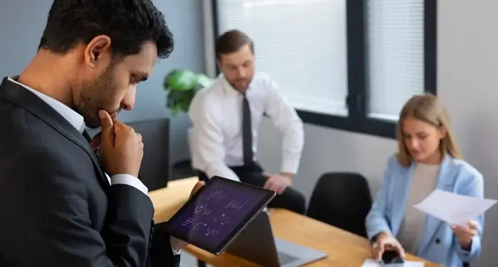 Empresario analizando gráficos financieros en una tableta digital mientras su equipo trabaja en la oficina.