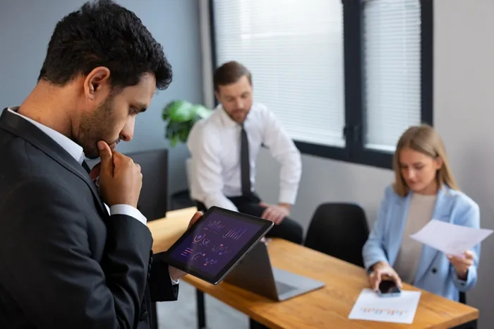 Empresario analizando gráficos financieros en una tableta digital mientras su equipo trabaja en la oficina.