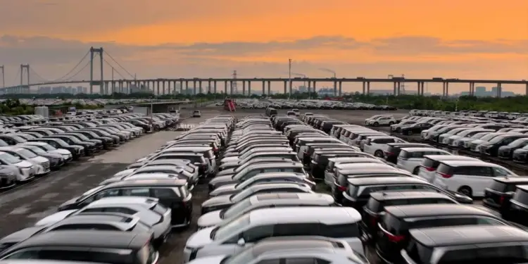 Amplio estacionamiento exterior con filas ordenadas de automóviles nuevos bajo un cielo al atardecer y un puente al fondo