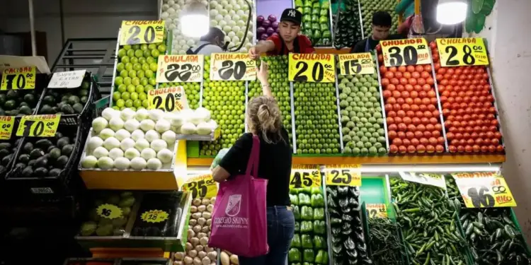 Puesto de mercado con abundancia de frutas y verduras frescas como limones, aguacates, cebollas y chiles. Se observan precios y clientes.