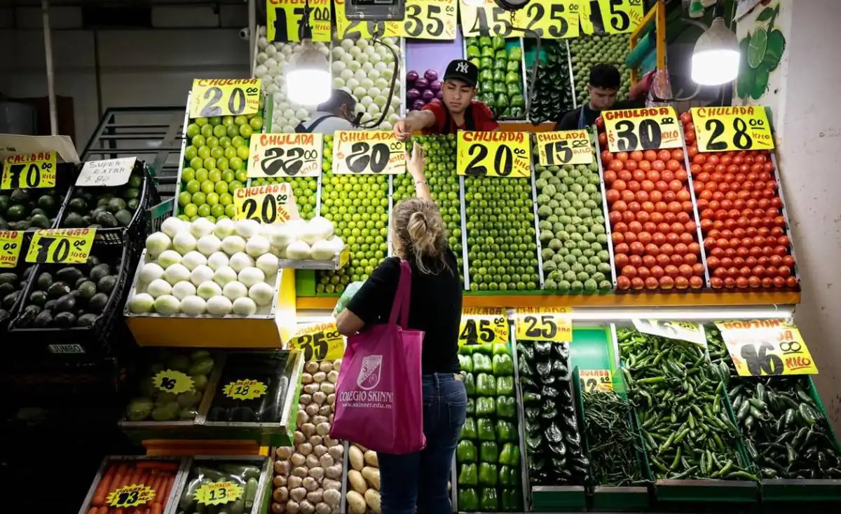 Puesto de mercado con abundancia de frutas y verduras frescas como limones, aguacates, cebollas y chiles. Se observan precios y clientes.
