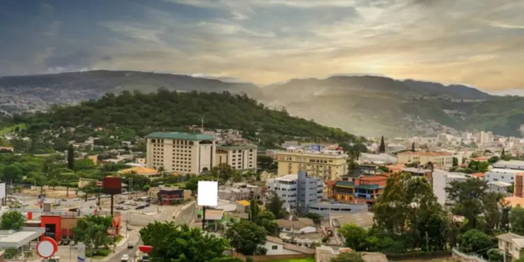 Vista aérea de una ciudad en un valle, con edificios comerciales, residenciales y carreteras. Montañas verdes rodean el área urbana bajo un cielo nublado.