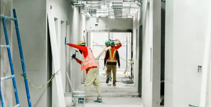 Obreros con cascos y chalecos reflectantes trabajando en un pasillo interior en construcción, instalando paredes y sistemas de techo.
