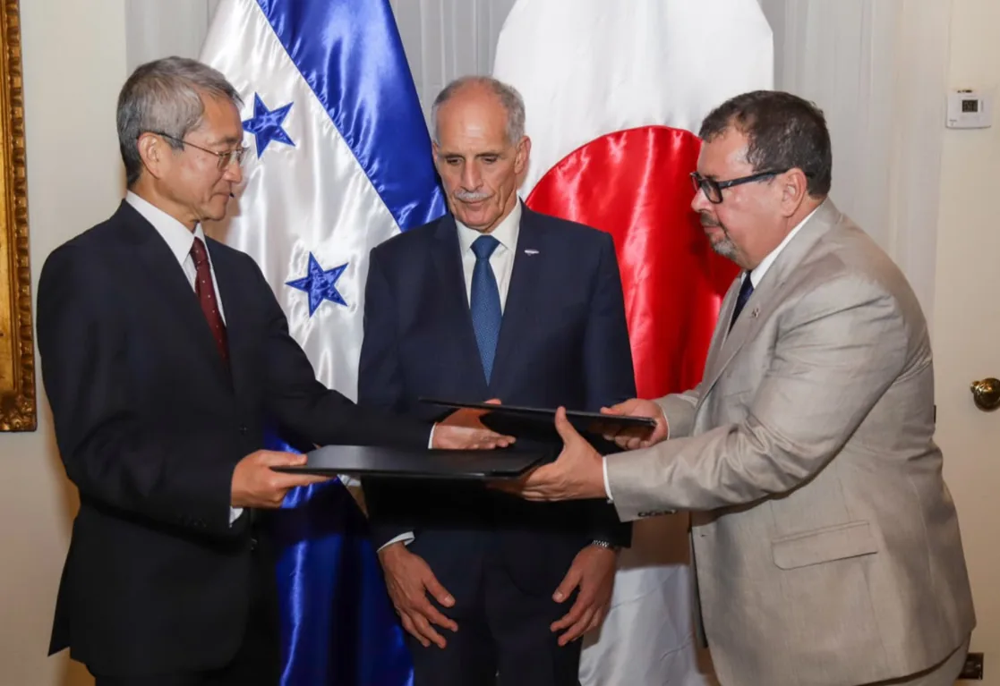 Tres hombres de negocios en trajes formales intercambian documentos frente a las banderas de Honduras y Japón, simbolizando un acuerdo bilateral.