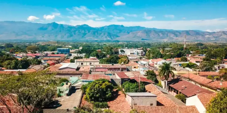 Vista panorámica de una ciudad con techos de tejas rojas rodeada de vegetación y montañas al fondo bajo un cielo azul.