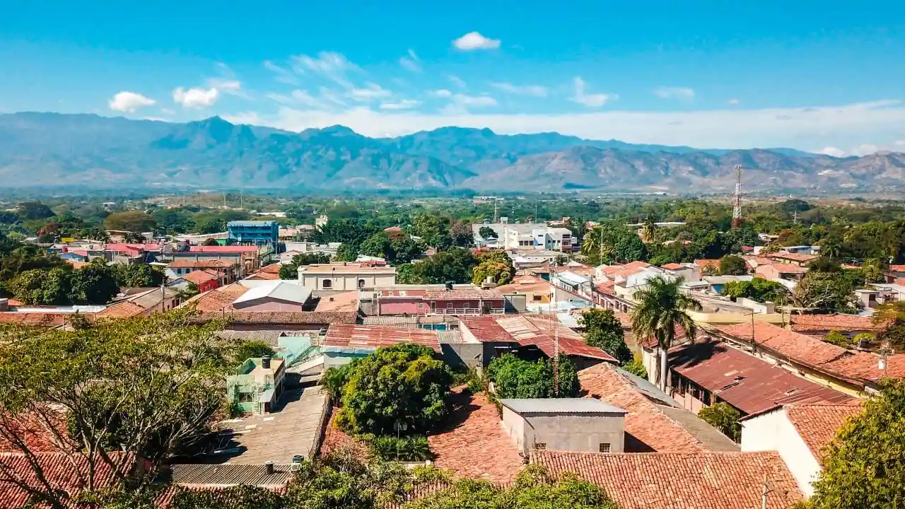 Vista panorámica de una ciudad con techos de tejas rojas rodeada de vegetación y montañas al fondo bajo un cielo azul.