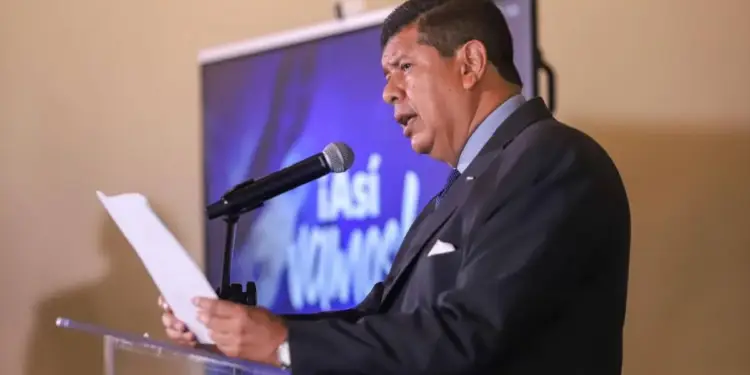 Hombre de mediana edad en traje oscuro y corbata azul, hablando en un podio con micrófono. Sostiene papeles y hay una pantalla con texto parcial detrás.