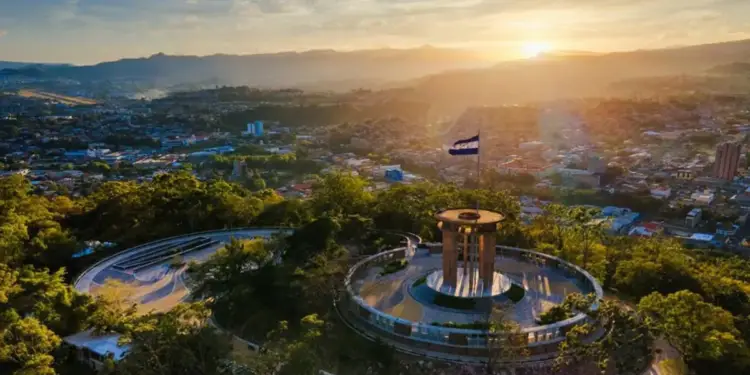 Vista aérea de una ciudad en un valle al atardecer, con un monumento circular y una bandera de El Salvador en primer plano. Montañas al fondo.