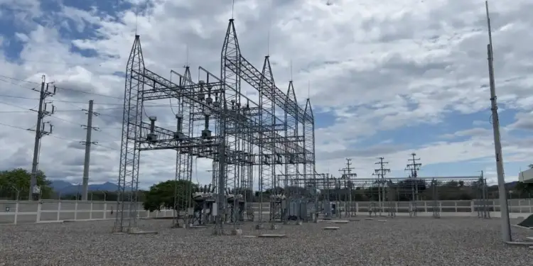 Vista panorámica de una subestación eléctrica con grandes estructuras metálicas, transformadores y líneas de alta tensión bajo un cielo nublado. El suelo es de grava y se aprecian montañas al fondo.