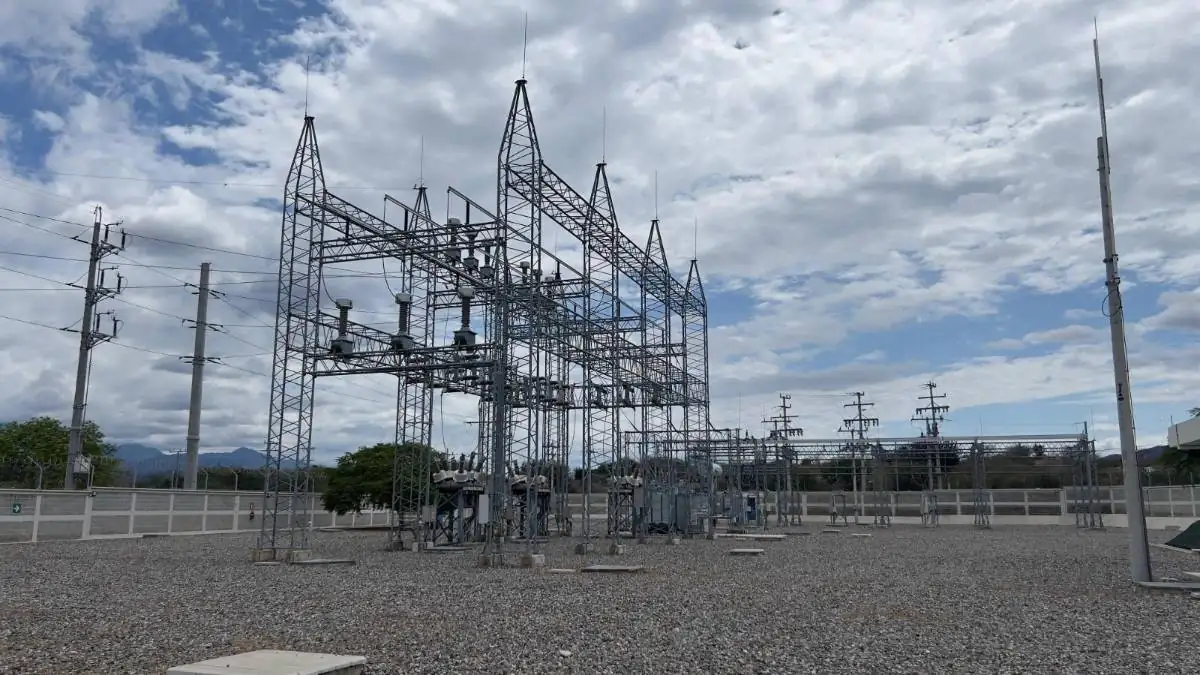 Vista panorámica de una subestación eléctrica con grandes estructuras metálicas, transformadores y líneas de alta tensión bajo un cielo nublado. El suelo es de grava y se aprecian montañas al fondo.
