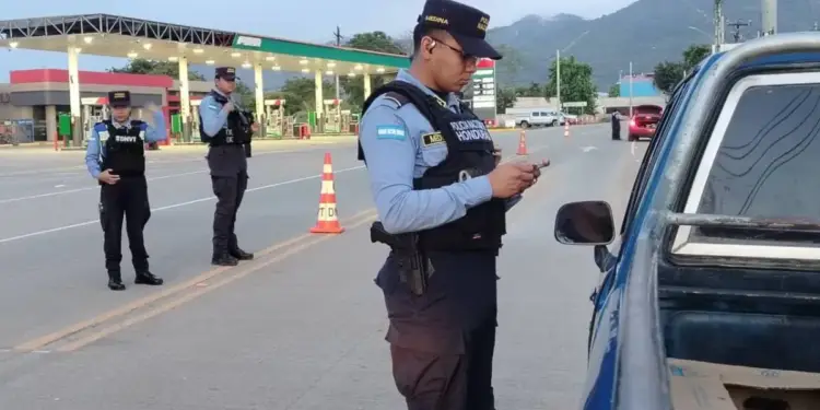 Oficiales de policía con uniforme azul y chaleco táctico realizando un control vehicular en una carretera cerca de una estación
