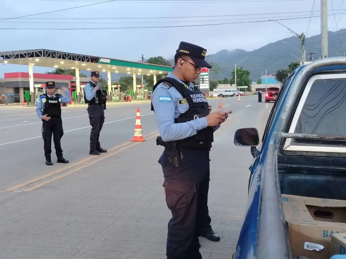 Oficiales de policía con uniforme azul y chaleco táctico realizando un control vehicular en una carretera cerca de una estación
