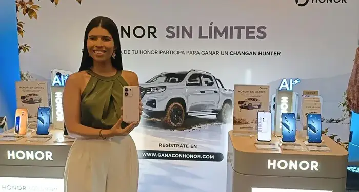 Mujer sonriente sosteniendo un teléfono móvil en un stand promocional de la marca HONOR con camioneta de fondo.
