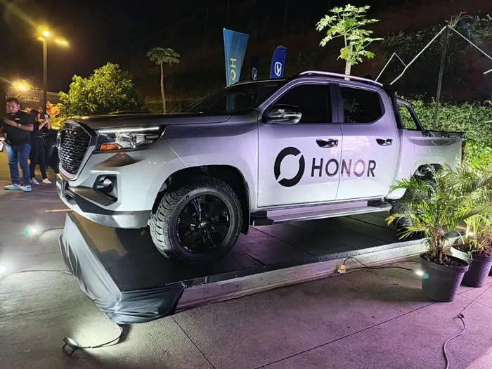 Camioneta pick-up plateada con la marca HONOR en la puerta, exhibida en una plataforma iluminada por la noche.