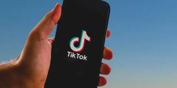 Mano sosteniendo un teléfono inteligente que muestra el logotipo de TikTok en la pantalla contra un fondo azul.