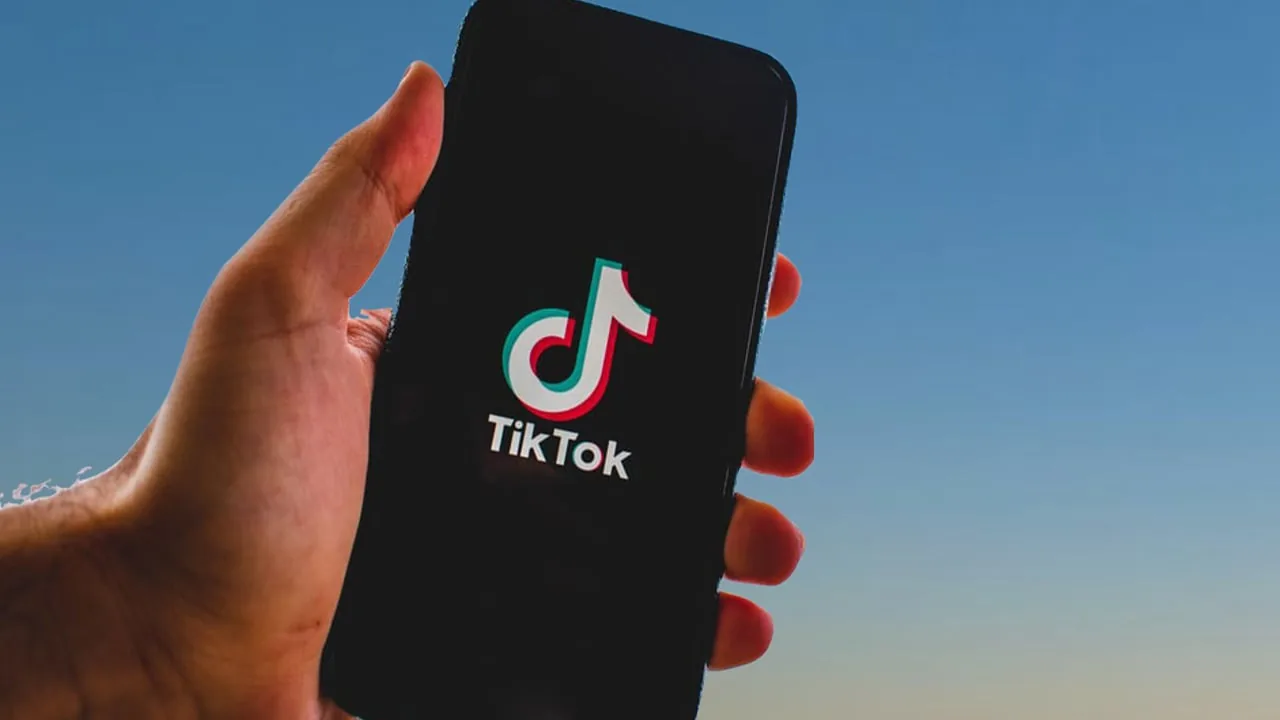 Mano sosteniendo un teléfono inteligente que muestra el logotipo de TikTok en la pantalla contra un fondo azul.