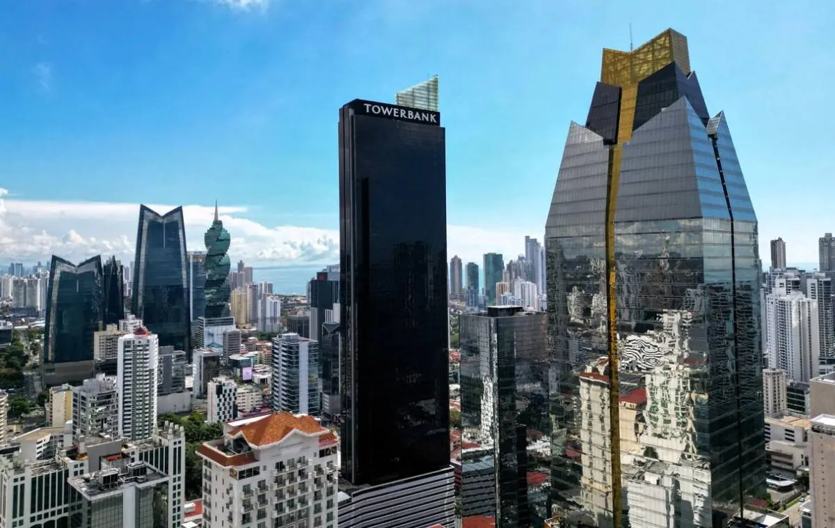 Vista aérea de un distrito financiero con rascacielos modernos, incluyendo el edificio Towerbank y otras estructuras de cristal y oro, bajo un cielo azul. Refleja dinamismo económico.