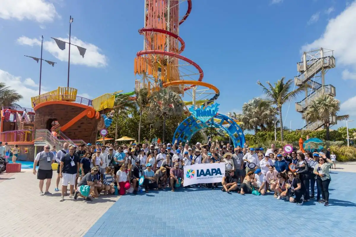 Gran grupo de profesionales de IAAPA América Latina y Caribe posando con un banner frente a atracciones de un parque acuático, incluyendo un barco pirata y toboganes coloridos, bajo un cielo azul.