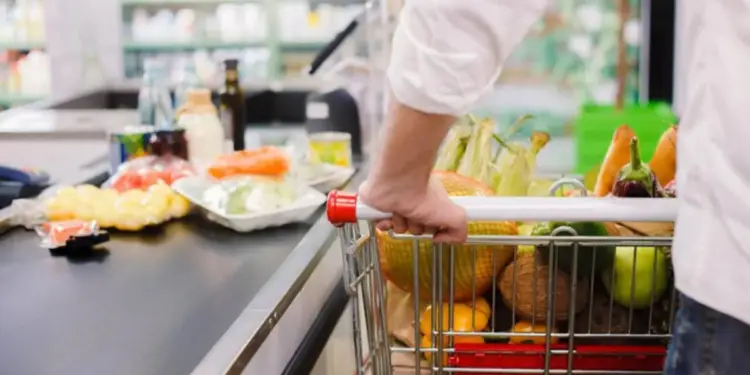 Un cliente empuja un carrito de supermercado repleto de productos frescos y envasados hacia la caja, con artículos ya en la cinta.