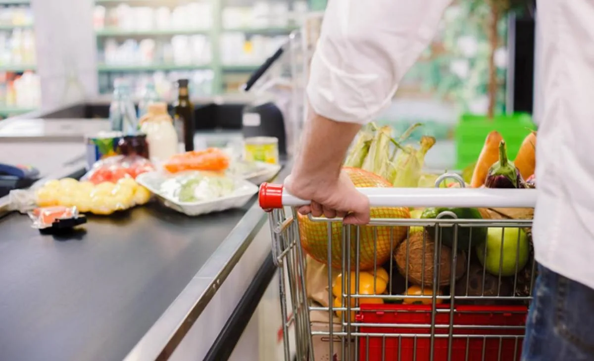 Un cliente empuja un carrito de supermercado repleto de productos frescos y envasados hacia la caja, con artículos ya en la cinta.