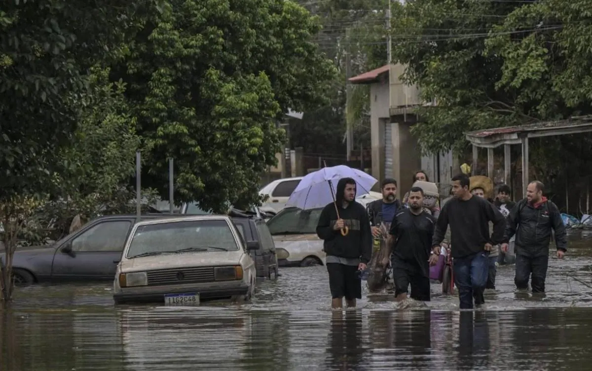Grupo de personas caminando por una calle residencial inundada con vehículos parcialmente sumergidos.