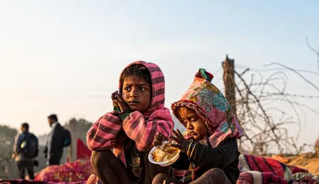 Dos niños sentados en el suelo, uno con las manos en la barbilla mirando a lo lejos, el otro comiendo de un plato. Ambos visten ropa abrigada y colorida.