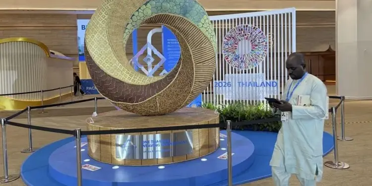 Escultura circular de gran tamaño con texturas y colores variados, representando "Tejiendo Naciones", ubicada en un evento financiero con un hombre revisando su teléfono móvil al lado.