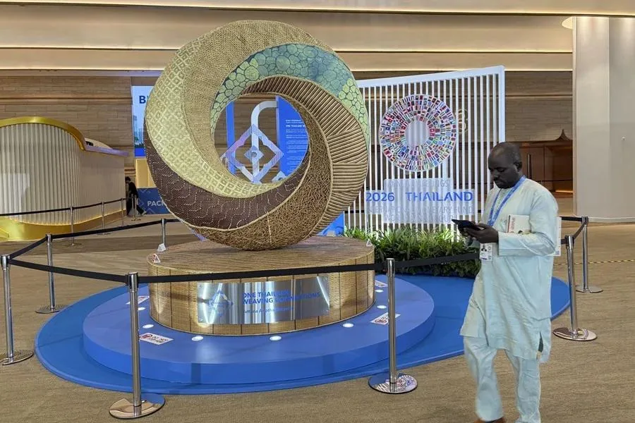 Escultura circular de gran tamaño con texturas y colores variados, representando "Tejiendo Naciones", ubicada en un evento financiero con un hombre revisando su teléfono móvil al lado.