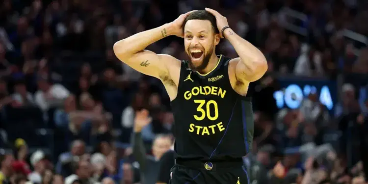 Un jugador de baloncesto con camiseta negra y amarilla 'GOLDEN STATE 30' celebra con las manos en la cabeza y expresión de alegría en un estadio lleno.