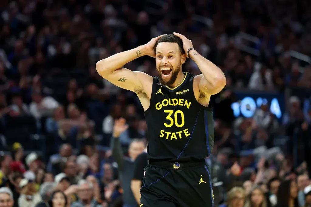 Un jugador de baloncesto con camiseta negra y amarilla 'GOLDEN STATE 30' celebra con las manos en la cabeza y expresión de alegría en un estadio lleno.