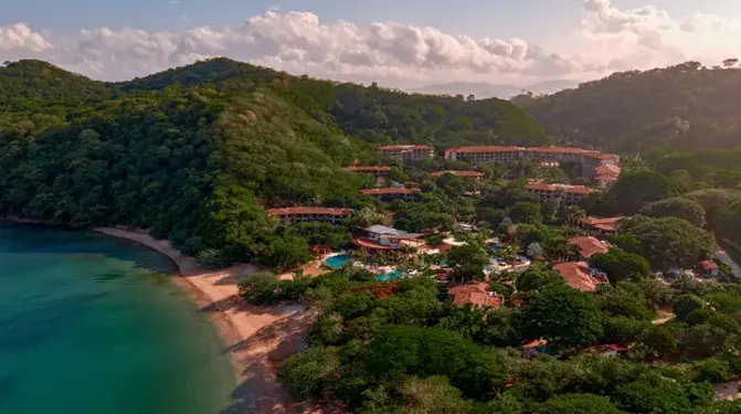 Vista aérea de un resort tropical con edificios de techos naranjas, piscinas y vegetación exuberante, junto a una playa de arena y el océano turquesa.
