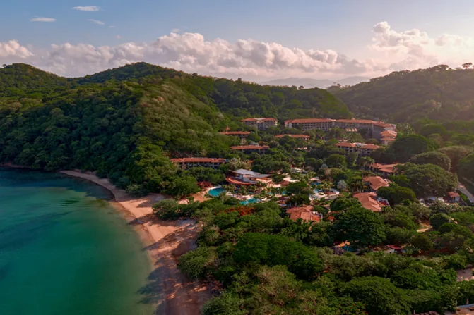 Vista aérea de un resort tropical con edificios de techos naranjas, piscinas y vegetación exuberante, junto a una playa de arena y el océano turquesa.