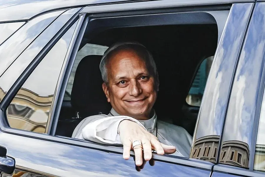 Hombre sonriente con vestimenta clerical asomado por la ventana de un coche oscuro. Reflejos de edificios y cielo en el cristal.