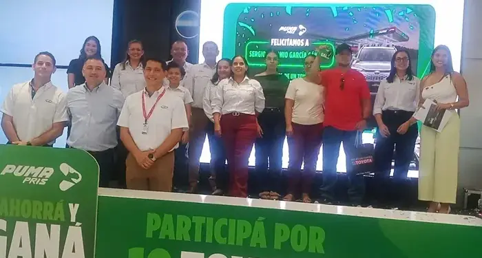 Grupo de personas sonrientes en un evento corporativo frente a una pantalla con un anuncio promocional.