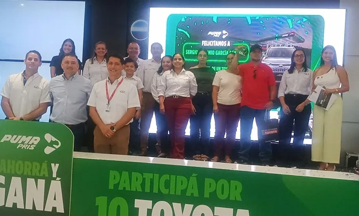 Grupo de personas sonrientes en un evento corporativo frente a una pantalla con un anuncio promocional.