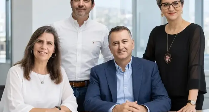 Cuatro profesionales, dos hombres y dos mujeres, posando en una oficina moderna. Dos sentados en una mesa de madera clara, dos de pie detrás. Ambiente de negocios.