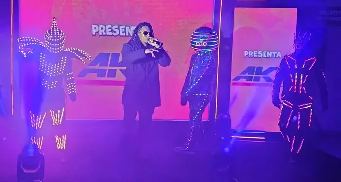 Un cantante en el centro con gafas de sol y rastas, acompañado por tres bailarines con trajes iluminados por luces LED en un escenario.
