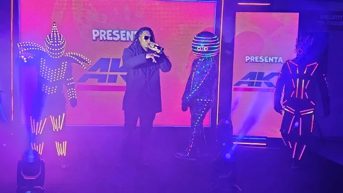 Un cantante en el centro con gafas de sol y rastas, acompañado por tres bailarines con trajes iluminados por luces LED en un escenario.
