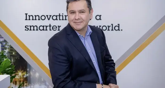 Hombre de negocios sonriente con traje oscuro y camisa azul en una oficina moderna.