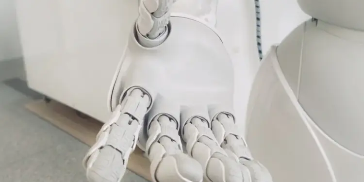 Primer plano de la mano blanca y articulada de un robot extendida hacia adelante en un entorno moderno.