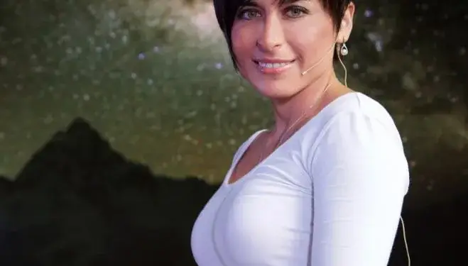 Mujer profesional de cabello corto y blusa blanca sosteniendo un dispositivo móvil frente a un fondo oscuro.