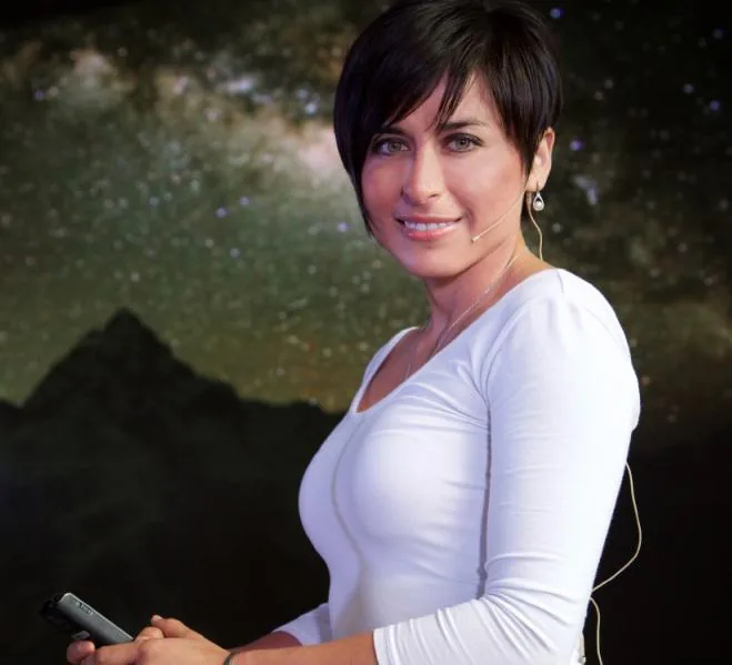 Mujer profesional de cabello corto y blusa blanca sosteniendo un dispositivo móvil frente a un fondo oscuro.