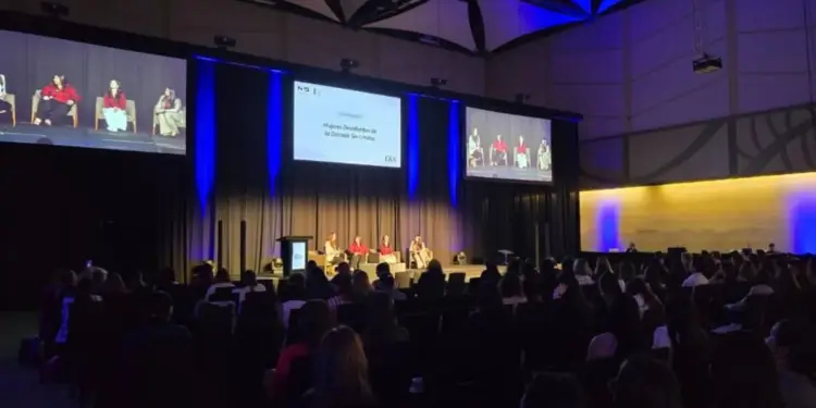 Tres mujeres sentadas en sillones en un escenario frente a una gran pantalla con texto de panel magistral